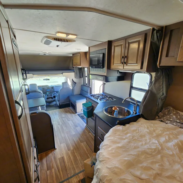 Interior of an RV rental, showing the kitchen with stove & sink, a bed with white bedding, and the front cockpit area.. Thor Majestic 2019