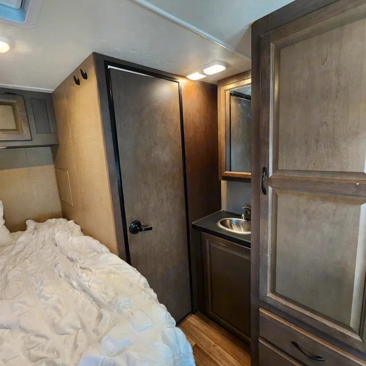 This RV rental offers a compact bedroom with white bedding, a functional vanity with sink, and dark wood cabinetry.. Thor Majestic 2019