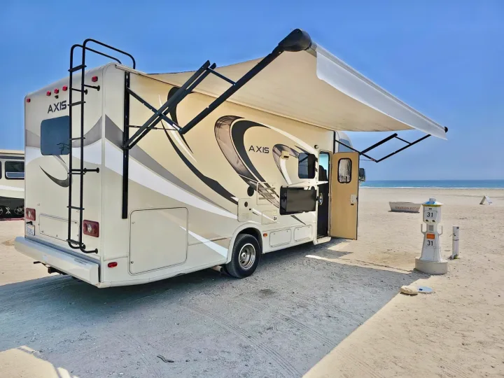 RV rental parked on a sunny sandy beach near the ocean, featuring an extended awning and outdoor TV.. Thor Axis 2017