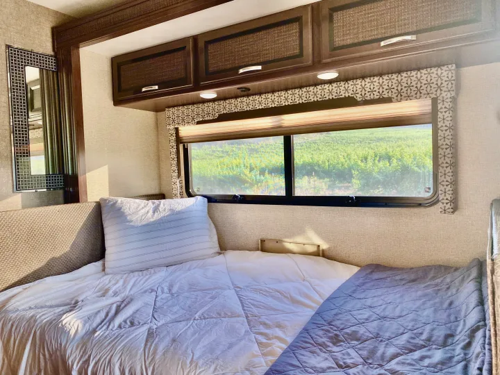 Cozy RV rental bedroom with a comfy bed, overhead cabinets, and a scenic window view of green vineyards.. Thor Axis 2017