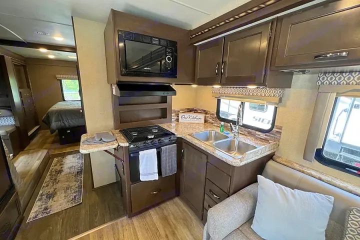 RV rental kitchen: a compact space with microwave, gas stove, double sink, brown cabinets, and counter space, visible next to a couch and bedroom.. Thor Motor Coach Freedom Elite 2017