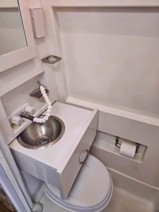Compact RV rental bathroom features a sink unit directly above the toilet, complete with a flexible faucet, mirror, and toilet paper holder.. Mercedes-Benz Sprinter 2005