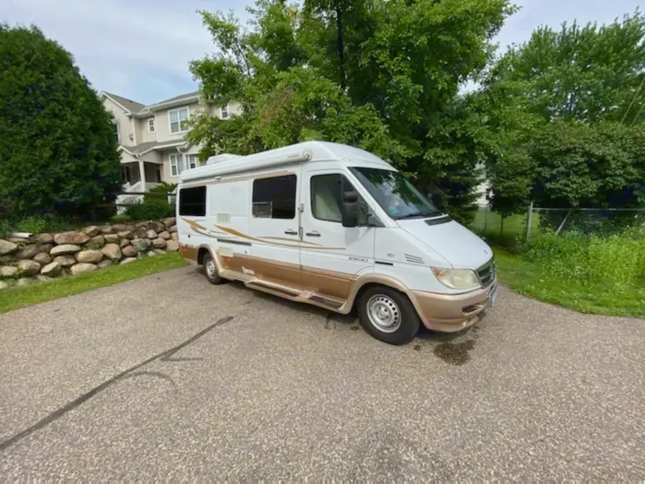 This white & gold Sprinter RV rental, ideal for a road trip or camping, is parked on a driveway with a house & trees.. Mercedes-Benz Sprinter 2005