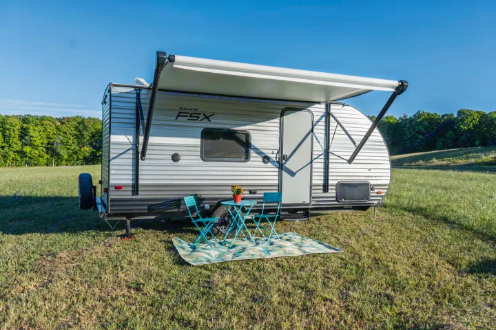 An RV rental travel trailer with an extended awning, outdoor patio set, and rug in a sunny, grassy field.. Forest River Salem Cruise Lite 2018