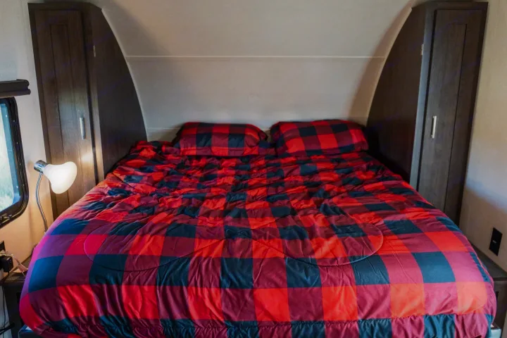 Inside an RV rental bedroom featuring a cozy bed with red and black buffalo plaid bedding, side cabinets, and a lamp.. Forest River Salem Cruise Lite 2018