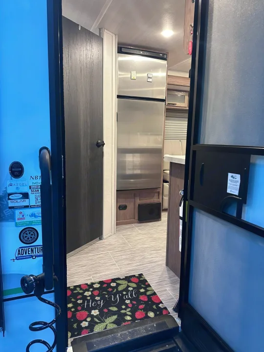 Interior view of an RV rental showing the kitchen area, fridge, and a "Hey Y'all" mat on the floor.. No Boundaries 19 2023