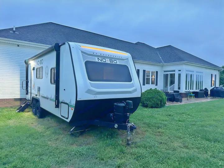 A white 'NO BO' travel trailer RV rental, parked on a green lawn at dusk beside a house. Ready for your outdoor adventure!. No Boundaries 19 2023