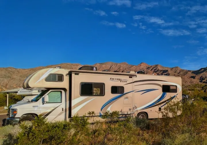 A beige RV rental in a vast desert landscape, nestled against rocky mountains under a bright blue sky. Ideal for nature travel.. Thor Freedom Elite 2018