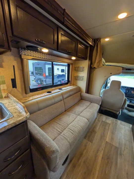 View inside an RV rental showing a tan sofa, dark wood cabinets, and kitchen area, with a window and driver's cab.. Thor Freedom Elite 2018