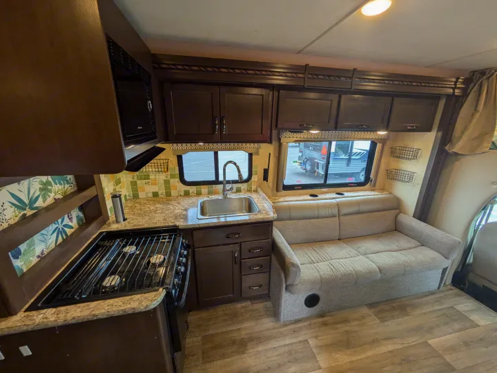 RV rental interior: A compact kitchen with stove, sink, microwave, brown cabinets, and a light-colored sofa for living space.. Thor Freedom Elite 2018