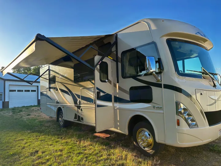 Light tan RV rental motorhome parked on green grass with awning extended. A white garage is visible behind it under a blue sky.. Thor A.C.E. 2017