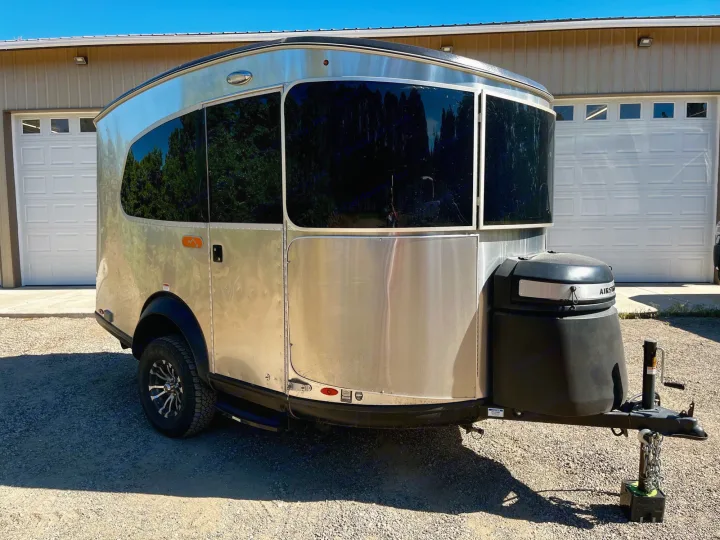 Sleek silver Airstream travel trailer for rv rental, parked on gravel in front of a building with garage doors.. Airstream Base Camp 2021