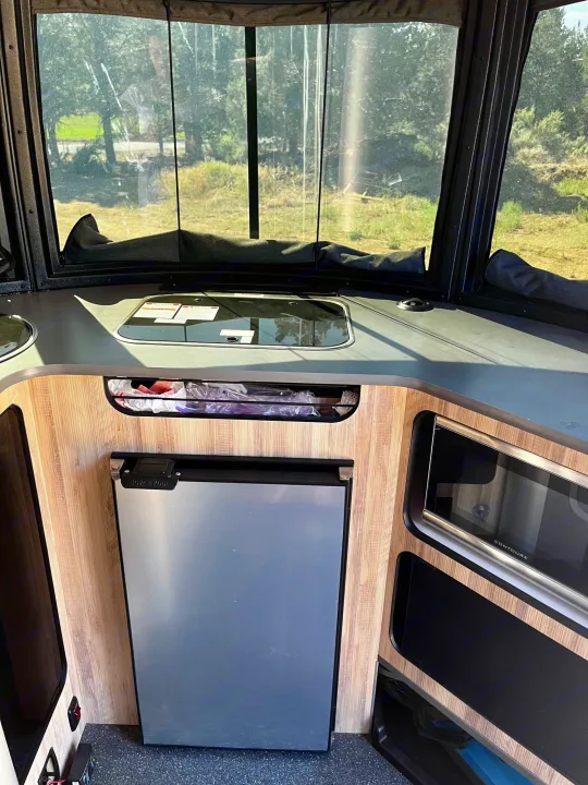 This RV rental shows its compact kitchenette with refrigerator, microwave, sink, and stovetop, offering a scenic window view.. Airstream Base Camp 2021
