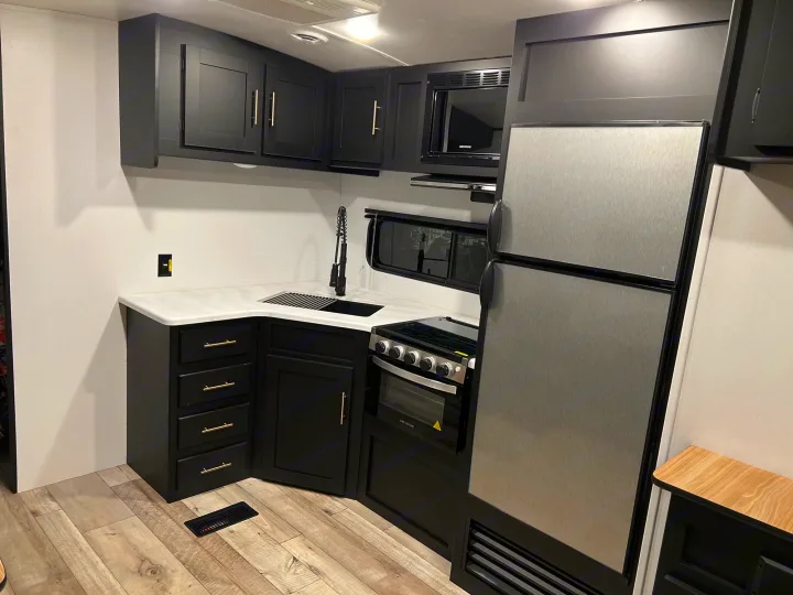Modern RV rental kitchen with sleek black cabinets, white counters, and stainless steel appliances, including a fridge, microwave, and stove.. Keystone RV Springdale 2022