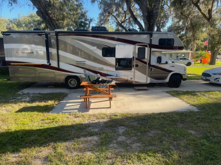 A cream and burgundy Sunseeker RV rental is parked at a campsite, with a small wooden picnic table on the concrete pad.. Forest River Sunseeker 2018