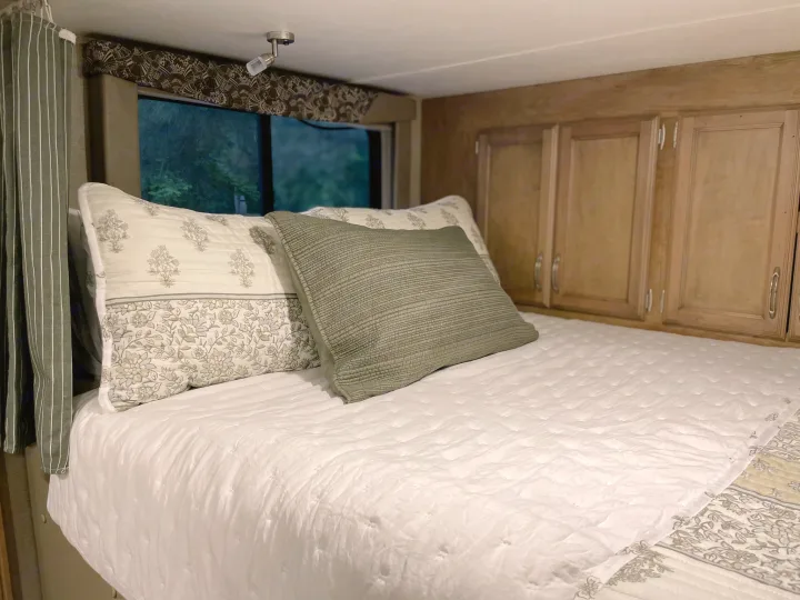 Cozy RV rental bedroom with a comfortable bed, patterned pillows, and a window view of lush trees, ideal for travel.. Gulf Stream Conquest 2017
