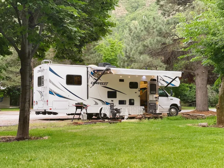 A white RV rental motorhome with an extended awning and string lights is set up at a shaded campsite, complete with a grill and chairs for outdoor relaxation.. Gulf Stream Conquest 2017