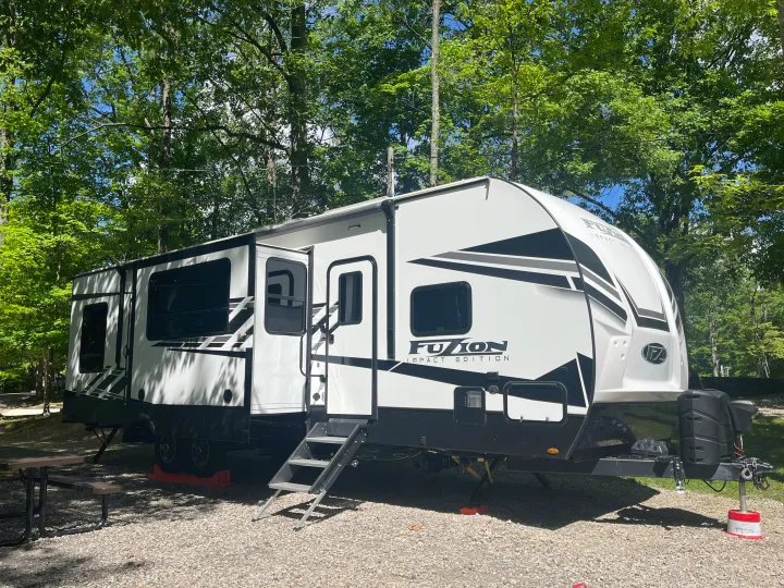 Modern white & black Fuzion travel trailer parked on gravel at a shaded campsite, ready for your next rv rental adventure.. Keystone RV Fuzion Impact Toy Hauler 2022
