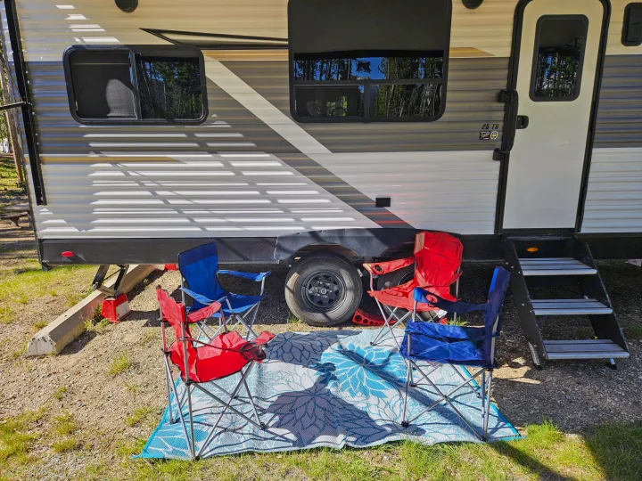 This outdoor rv rental setup features a large RV, multiple red and blue camping chairs, and a decorative mat at a sunny campsite.. Open Range Open Range 2023