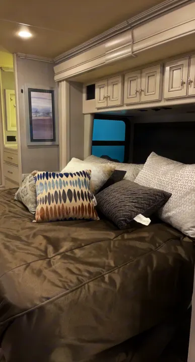 A modern RV rental bedroom featuring a made bed with multiple pillows, overhead cabinets, and a framed picture.. Holiday Rambler Nautica 2022