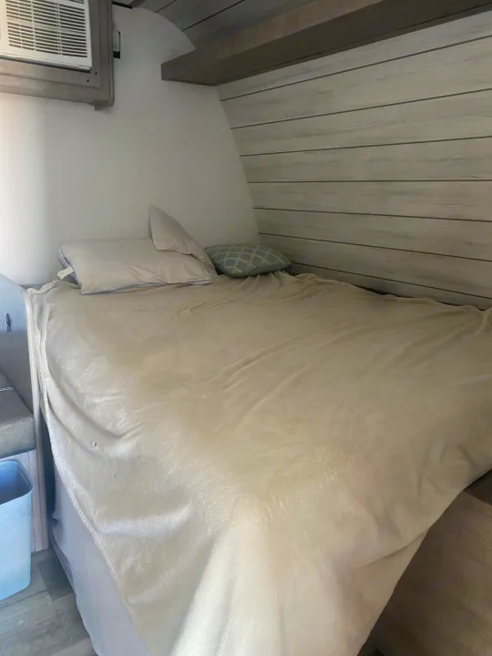 Inside an RV rental: a compact sleeping area with a bed, light blanket, pillows, and wall-mounted AC unit, offering cozy comfort.. Coleman Lantern LT 17b 2024