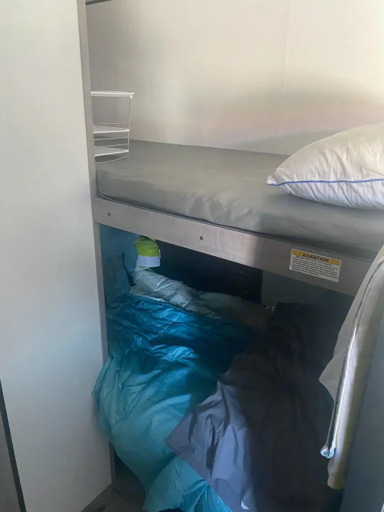 RV rental bunk beds: The upper bunk features a mattress and pillow, the lower bunk holds bedding, with a small storage rack.. Coleman Lantern LT 17b 2024