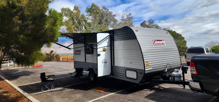This Coleman travel trailer rv rental is set up in a sunny parking spot with an awning extended, chairs out, and a truck hitched. Mountains are in the background.. Colemen Dutchmen 17R 2024