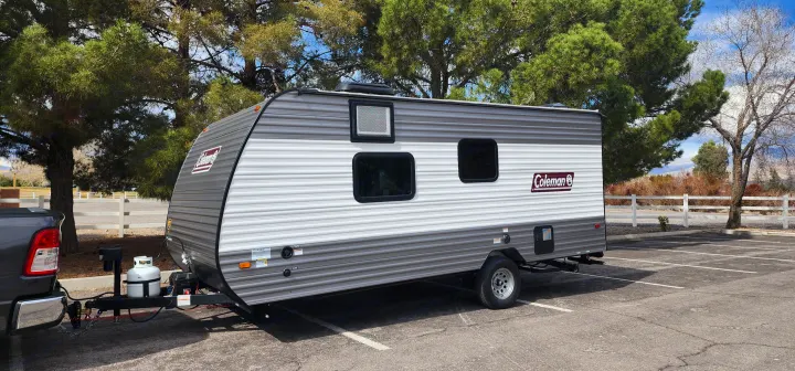 A modern gray and white Coleman travel trailer, ready for rv rental, is parked and hitched to a pickup truck outdoors.. Colemen Dutchmen 17R 2024