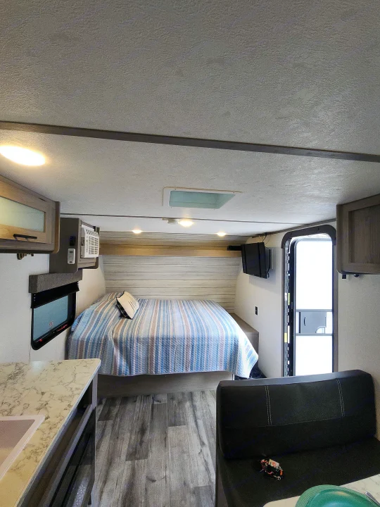 RV rental interior showcasing a cozy bedroom with a striped quilt, compact kitchen, and modern wood-look flooring.. Colemen Dutchmen 17R 2024