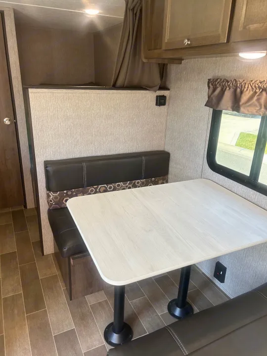 RV rental interior: dinette with light wood table and dark benches, an upper bunk bed, and overhead cabinets.. Coleman Lantern 17FQ 2020