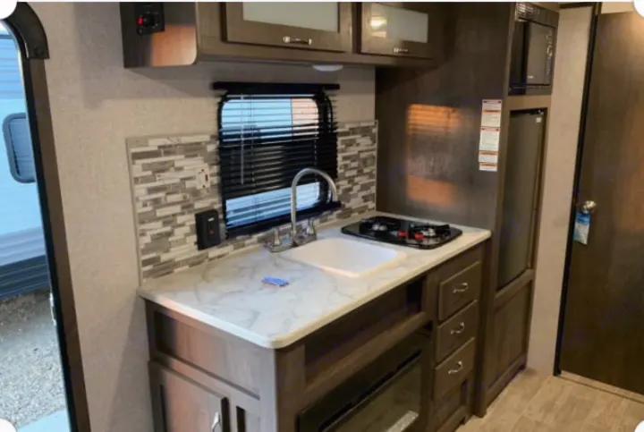 Compact RV rental kitchen featuring a sink, two-burner stove, microwave, fridge, and backsplash for your travels.. Dutchmen Coleman Lantern 2021