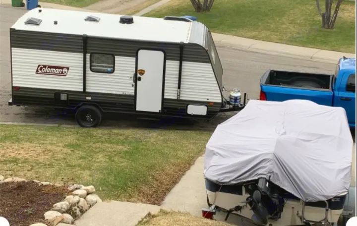 An RV rental, a black and white travel trailer, is parked with a blue truck and a covered boat in a residential area.. Dutchmen Coleman Lantern 2021