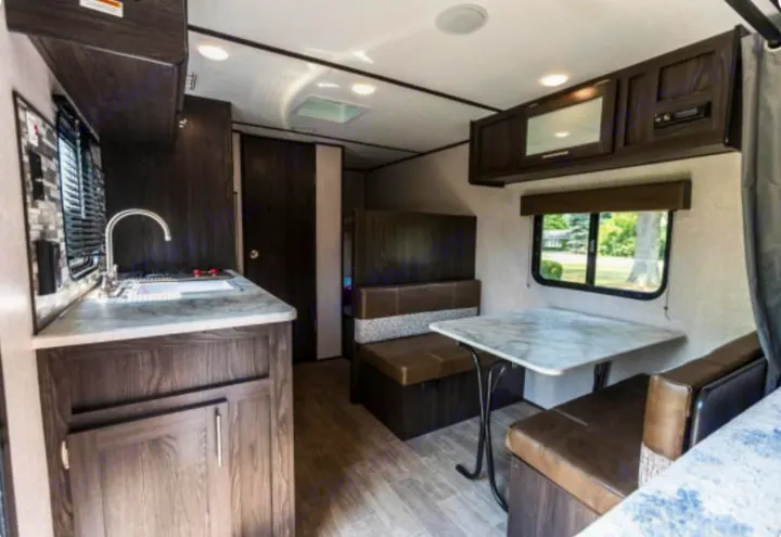 Inside a stylish rv rental: dark wood kitchen cabinets, light countertops, and a cozy dinette with brown seating.. Dutchmen Coleman Lantern 2021