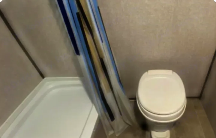 Compact RV rental bathroom with a white toilet and a small shower. A blue, black, and tan striped curtain hangs by the shower.. Dutchmen Coleman Lantern 2021