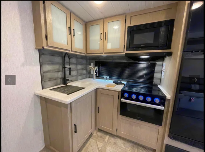 A sleek RV rental kitchen showcases light wood cabinets, black appliances, sink, range, and a distinctive gray tiled backsplash.. Forest River Wildwood Platinum 2024