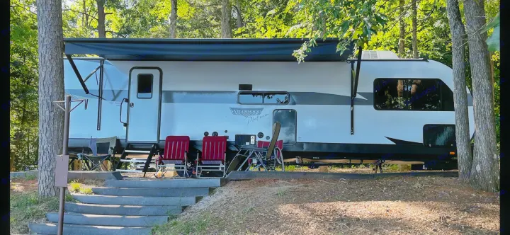 Wildwood Platinum RV rental at a shaded campsite, awning extended, with outdoor chairs by the steps. Perfect for leisure.. Forest River Wildwood Platinum 2024