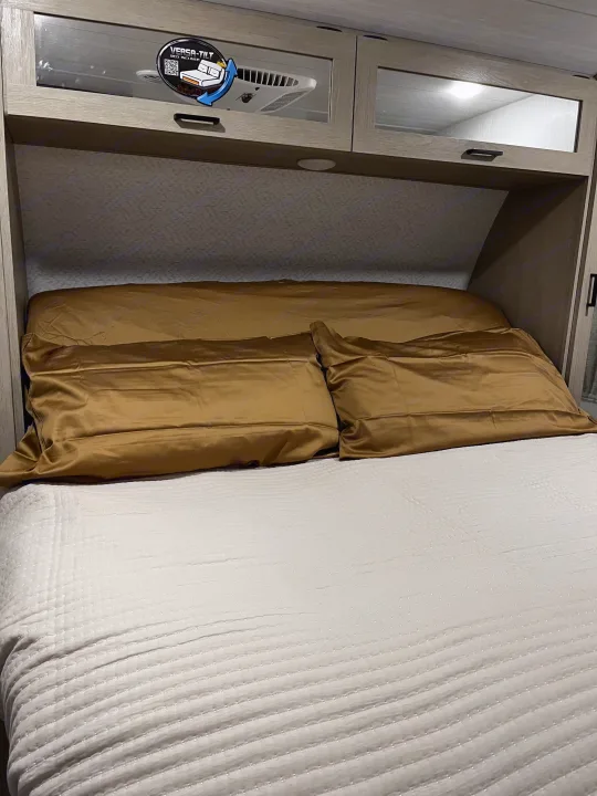Bright RV rental bedroom showcasing a comfortable bed with golden pillows, sleek overhead cabinets, and an adjustable bed feature.. Forest River Wildwood Platinum 2024