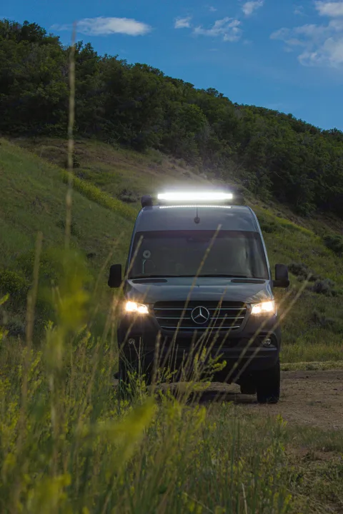 Dark Mercedes Sprinter RV rental, lights on, parked on a dirt path through tall grass & hills at dusk.. Mercedes-Benz Sprinter 4x4 2022