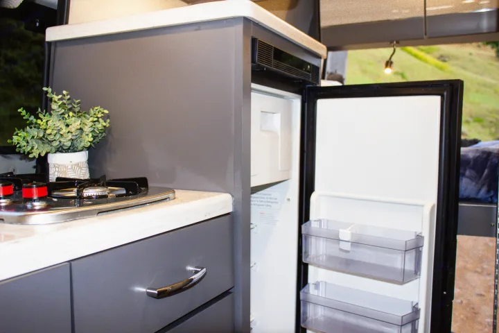 Inside an RV rental: a modern, compact kitchen with a gas stove, open refrigerator, and light grey cabinetry.. Mercedes-Benz Sprinter 4x4 2022