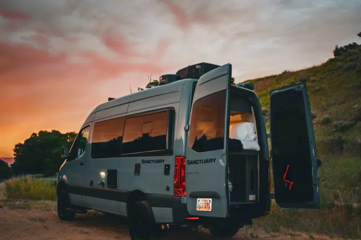 Gray camper van RV rental parked on a dirt road at sunset, rear doors open revealing the cozy interior. Ready for adventure!. Mercedes-Benz Sprinter 4x4 2022