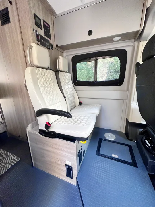 Inside a modern RV rental: white quilted seating, various control panels, an overhead cabinet, and blue patterned flooring.. Winnebago Solis 2022
