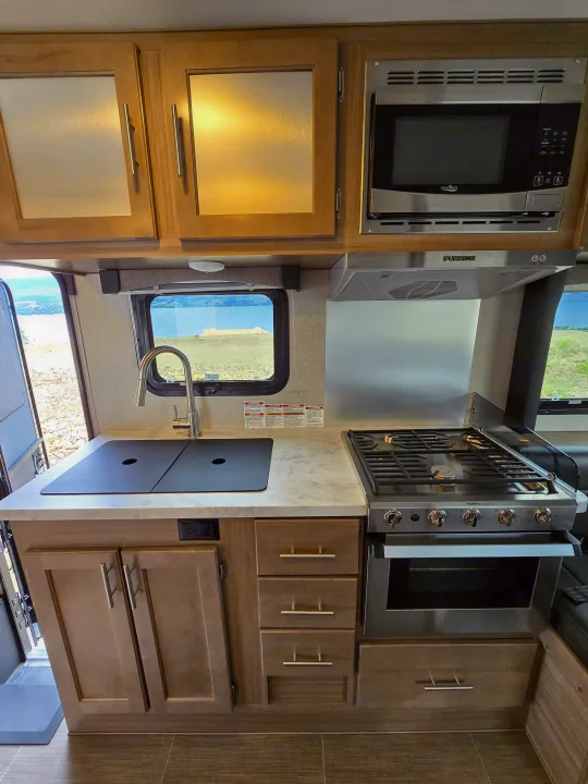Modern RV rental kitchen with a stainless steel stove, microwave, sink, wood cabinets, and scenic window views.. ALP Adventurer 19RD 2021