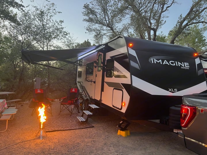 Relaxing by a cozy campfire with an Imagine XLS RV rental at dusk. A perfect campsite setup for outdoor adventures.. Grand Design Imagine XLS 2024