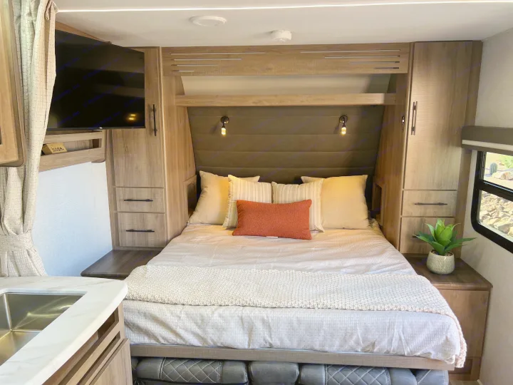 Cozy RV rental bedroom with a comfortable bed, mounted TV, built-in storage, and partial kitchen view. Ideal for trips.. Grand Design Imagine XLS 2024