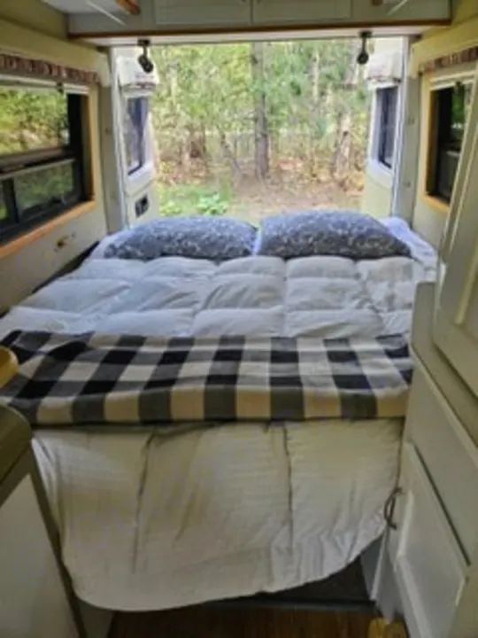 Comfortable RV rental bedroom: A cozy bed with white comforter, gray pillows, plaid blanket, and a serene wooded view.. Mercedes-Benz Sprinter 2005