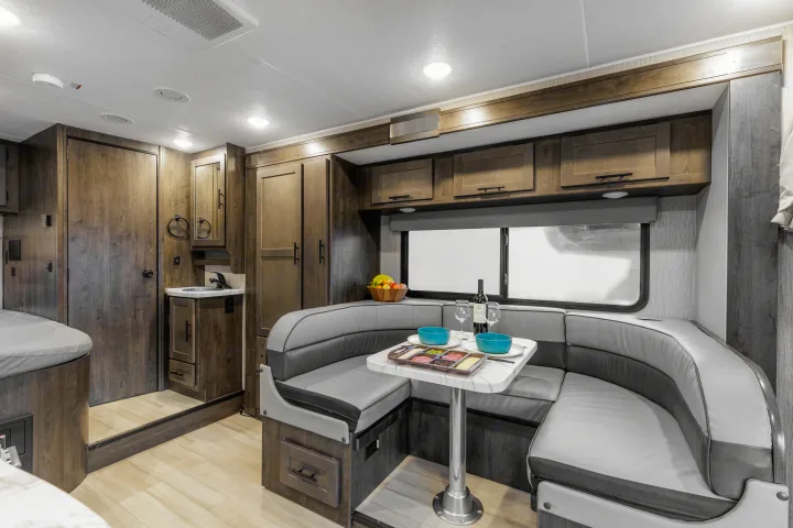 Inside an RV rental: a modern wood interior with a gray U-shaped dinette, dining table set, and compact kitchen.. Coachmen Prism 2024