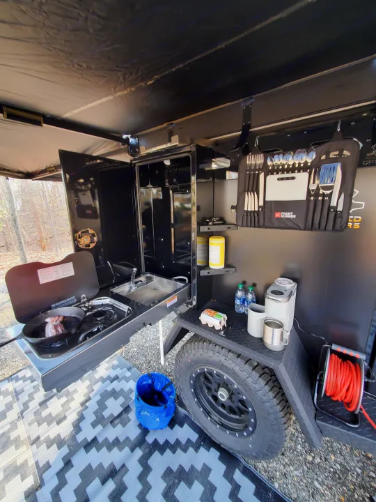RV rental outdoor kitchen. Cooking bacon, sink, coffee maker, and organized utensils provide a complete camping setup.. Off Grid Trailers Off Grid Trailers Trailer 2021