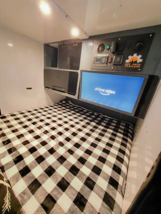 Cozy RV rental interior: a comfortable bed with a checkered blanket, TV showing Prime Video, and an off-grid control panel.. Off Grid Trailers Off Grid Trailers Trailer 2021