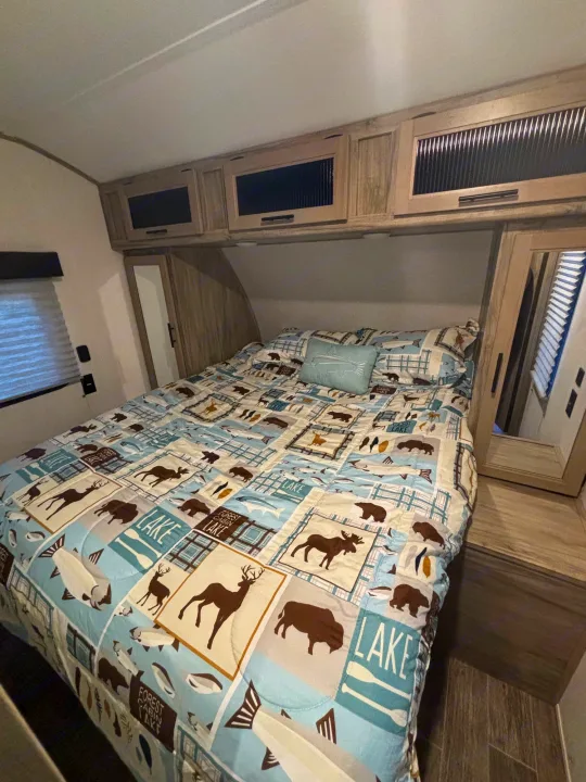 RV rental bedroom: a cozy retreat with nature-patterned bedding, overhead storage, and window blind.. Forest River Cherokee Alpha Wolf 2021