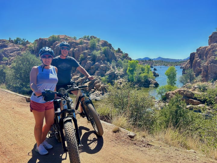 Two people enjoy an electric bike ride by a beautiful lake with unique rock formations, ideal for an RV rental getaway!. Grand Design Imagine XLS 2024